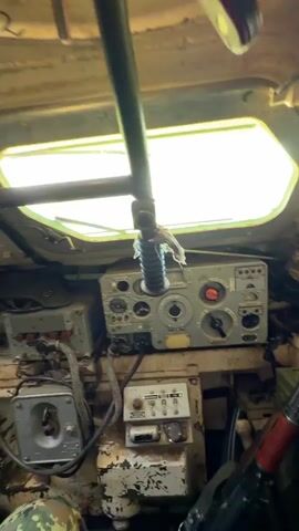 This is a first-person perspective video from inside a military vehicle, likely a tank. The camera pans across the interior, showing various mechanical components, controls, and equipment. In the first few seconds, we see a dashboard with numerous gauges and switches, as well as a steering column. A person's legs, wearing camouflage pants and black boots, are visible, suggesting they are operating or navigating the vehicle. The interior appears worn and functional, with exposed wiring and metal surfaces. Later in the video, a rifle with a red wooden grip is seen resting against the interior wall. The view occasionally shifts to the outside through the vehicle's windows, revealing a bright, possibly sunlit environment with green foliage. The overall impression is of a close-up, inside view of a military vehicle's operational environment.