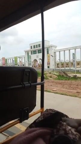 The video shows a view from inside a three-wheeled vehicle, likely a tuk-tuk. The camera is pointed forward and slightly to the right. In the foreground, the dark brown seat of the vehicle is visible, along with a black metal bar. A patterned fabric is draped over the seat in the lower right corner. The main focus of the video is a large, modern white building with green window frames and a prominent arched entrance. A long colonnade stretches out from the building to the left, also white with vertical elements. Several colorful flags or banners are displayed along the colonnade and in front of the building, some featuring green designs. The ground in front of the building appears to be a paved area and some undeveloped land with scattered debris. The sky is overcast. The vehicle is moving, causing a slight blur to the background.