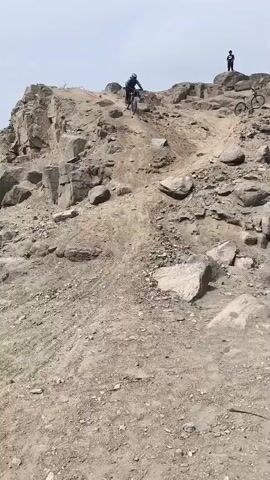 A mountain biker in a dark green and black kit attempts to descend a very steep, rocky, and dusty hill in what appears to be an arid environment. The rider navigates a section covered with large and small rocks, but loses balance and is dismounted from their bike. The bike tumbles a short distance down the slope. The rider quickly recovers, gets to their feet, and goes to retrieve the fallen bicycle. Two other individuals and another mountain bike are visible further up the hill, observing the descent. The sky is bright but overcast or hazy, and the terrain is dry, typical of a desert landscape near Trujillo, Peru. No discernible text is visible.