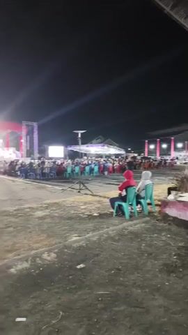 A night-time outdoor event is captured, with a large crowd gathered to watch a performance on a brightly lit stage. The stage is adorned with red and white banners and features several figures on it. A screen is also visible to the left of the stage. People are seated in rows of chairs, arranged in front of the stage and extending outwards. Several individuals are standing, and some are walking through the crowd. The ground appears to be dirt or gravel. In the foreground, two people are seated in green chairs, facing away from the camera and towards the event. A parked motorcycle is partially visible in the lower left corner. The sky is dark, indicating it is nighttime.