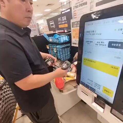 A person is using a self-checkout machine in what appears to be a grocery store. The person is holding a scanner and scanning items. The self-checkout machine has a touch screen displaying a receipt with various items and their prices. There are baskets of merchandise behind the person. The environment is brightly lit, typical of a retail setting.
