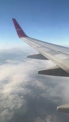 The video shows a view from an airplane window. The wing of the airplane is visible in the foreground, with a red tip featuring a white logo. Below the wing, there are dense white and gray clouds stretching to the horizon. The sky above is a clear, bright blue. The camera angle suggests the plane is in flight.