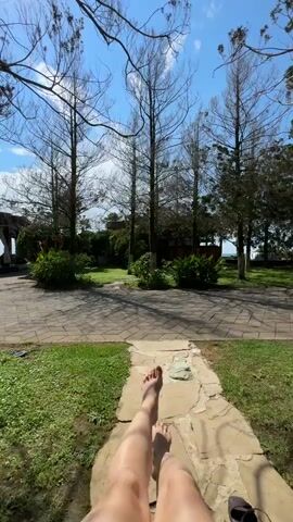 This video shows a first-person perspective of someone relaxing outdoors. The subject's bare feet and legs are visible in the foreground, resting on a light-colored, paved pathway. A black flip-flop lies nearby on the same path. The path leads into a courtyard area paved with interlocking stones, surrounded by lush green grass. Tall, bare trees with thin branches dominate the mid-ground, their stark silhouettes contrasting with the bright blue sky above. Behind the trees, hints of foliage and buildings can be seen, suggesting a developed area. The lighting and bright sky indicate it is daytime, likely sunny and warm. The overall mood is one of relaxation and leisure. The location context provided is Chakvi, Georgia, which is a coastal town known for its subtropical climate and beaches.