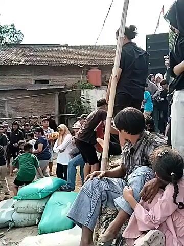A group of people are gathered outdoors in what appears to be a village setting. In the foreground, a person in a black headscarf and white pants stands holding a phone. To their left, a person wearing a plaid shirt and jeans sits on a green object, looking towards the center of the scene. In the center, two individuals in black clothing are engaged in an activity that involves balancing on what looks like a large, flat object, possibly a raft or a decorative piece. One of them is being supported by another person in a black shirt. A crowd of onlookers, including women, men, and children, are observing the activity from behind. In the background, traditional houses with tiled roofs are visible, along with some trees and a large speaker system mounted on a pole. The ground is uneven and appears to be dirt or gravel. The overall atmosphere seems to be one of community gathering and performance.