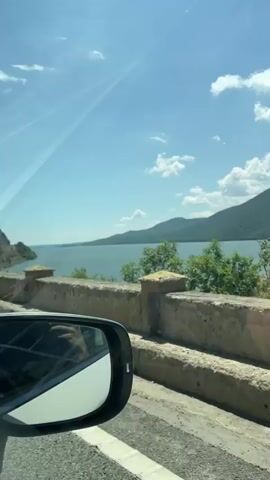 The camera is inside a moving vehicle, recording a view from the window as the vehicle drives alongside a large body of water. Lush green mountains and trees are visible on the far side of the water, under a bright blue sky with scattered white clouds. A road barrier is visible outside the window, and at 0:00, a blue parking sign with "1000m" is visible on a pole. The reflection of a car mirror is visible in the bottom left corner of the frame.