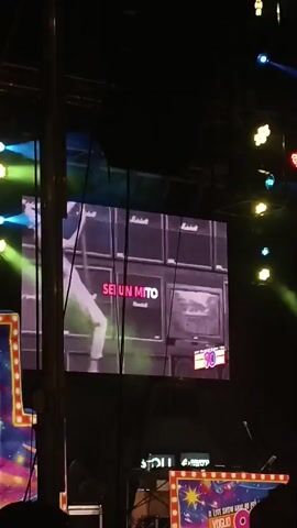 A video shows a large screen displaying a performance. The screen shows a person hanging from a trapeze, with text that reads "SEI UN MITO". The screen then transitions to show close-ups of three faces, with text that reads "SEI UN MITO PER ME". The background of the screen shows a stack of amplifiers and a logo with the number "90". The video appears to be taken from an audience's perspective at an outdoor event, with stage lighting and scaffolding visible. The bottom of the screen has a banner with colorful lights and text that says "90" and "LIVE SHOW ANNI '90 VOGLIO TORNARE".