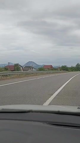 The video shows a view from inside a vehicle driving on a road. The road is paved with asphalt and has white lane markings. On the left side of the road, there is a metal guardrail and beyond that, some houses and trees. In the distance, there is a mountain. On the right side of the road, there is a truck pulling a flatbed trailer with red sides. Further down the road, there are a few other vehicles. The sky is overcast with clouds. The footage appears to be taken during the daytime.