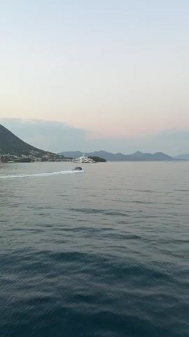 A jet ski with blue lights speeds across the water, leaving a white wake. In the distance, a large white yacht is anchored near a coastal town and hills. The camera then pans to show people on a boat, all dressed in white, looking out at the water and the receding jet ski. The sky is a pale, dusky blue.