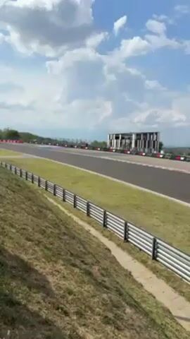 The video shows a racetrack on a sunny day with a partly cloudy sky. The camera is positioned on a grassy hill overlooking a straight section of the track. A long row of metal barriers lines the bottom of the hill, separating it from the track.  Two Formula 1 cars race down the track, followed by a third. The architecture of the race complex can be seen in the distance.