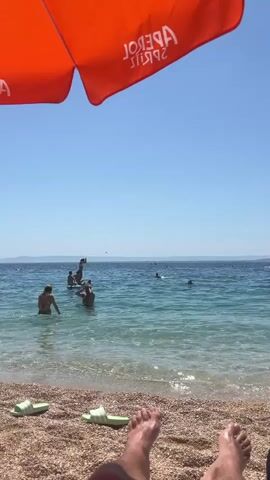 A first-person perspective video of a beach scene. The foreground shows a person's bare feet and legs resting on a pebbly beach, with two green slides nearby. An orange umbrella with the white text "APEROL SPRITZ" is visible at the top of the frame. The middle ground features the turquoise ocean with several people swimming and playing in the water. Some individuals are on paddleboards. The background shows a hazy coastline under a clear blue sky. The overall ambiance is that of a sunny day at the beach.