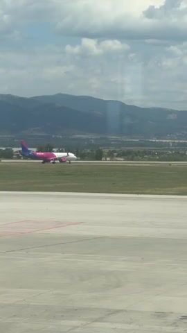 A wizz air airplane with pink and white livery is taxiing on a grassy area adjacent to an airport runway. In the foreground is a concrete surface with red markings. In the background, there are trees, hills, and mountains under a cloudy sky. The airplane is moving from left to right across the frame. The camera is stationary and facing the airplane.