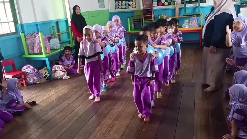 A group of young children, some wearing purple and white uniforms and headscarves, participate in an activity in a room that appears to be a classroom or play area. The room has wooden floors and colorful walls with decorations. In the foreground, several children are lined up, each holding a large blue balloon between their knees or with their hands. They are walking forward as a group, forming a train-like formation. Some children in the back are looking ahead, while others in the front are focused on the task.

To the left of the main line of children, a group of younger children are seated on the floor, observing the activity. One child is holding a phone and appears to be taking a photo or video.

In the foreground, a child in the purple and white uniform is bending down and interacting with cups placed on a wooden surface, possibly a table or desk. They are holding two cups, one of which is stacked on top of another.

Two adult women are present in the scene. One woman, wearing a hijab, is standing on the right side of the frame, appearing to observe or guide the children. Another woman, also wearing a hijab, is standing on the far left, partially visible.

The overall atmosphere appears to be one of playful activity and learning, with children engaged in a coordinated game. The room is brightly lit, suggesting it is daytime.