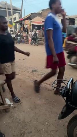 A woman walks down a dirt road in a marketplace. She is wearing a black t-shirt and tan shorts, with grey slippers. She carries a black plastic bag in her right hand. Behind her, a yellow motorized tricycle is visible. Several other people and motorcycles are present in the background. The area appears to be an open-air market with stalls selling goods. The road is unpaved and dusty. The lighting suggests it is daytime.