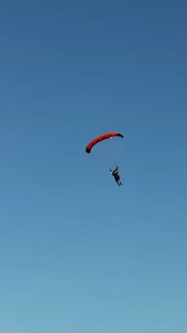 A tandem skydiver with a red parachute descends against a clear blue sky. The skydiver, wearing dark clothing and a helmet, is suspended below the parachute by lines. As the skydiver gets closer to the ground, a structure that appears to be a picnic table comes into view, and then a small building with a sign that has the letter "B" on it. The ground appears to be a grassy field with trees in the distance.