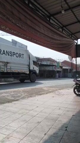 The video shows a street scene from under a canopy. A large white truck with "ROYAL POWER TRANSPORT" written on its side is moving from left to right. A motorcycle is parked on the right side of the frame, facing left. Further down the street, another truck, this one yellow with blue and red stripes, is also moving from left to right. A person is riding a motorcycle, moving from left to right in front of the buildings. The environment appears to be a commercial area with buildings lining the street. The ground is paved with concrete tiles in the foreground and asphalt further away. The overall lighting suggests daytime.
