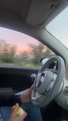 The video captures an internal view of a car, facing forward towards the driver and the road ahead, set in Sidi Abdallah Ghiat, Morocco. A person is seated in the driver's seat, dressed in a dark long-sleeved top and light blue ripped jeans, with a white face mask covering their nose and mouth. Their left hand rests on the steering wheel, which is light grey with a central emblem, indicative of a Fiat 500 interior. In their right hand, they hold a piece of light-colored fruit, possibly an apple or pear. Their right foot, clad in a textured, light-colored shoe, is visible resting on the car floor.

The vehicle is in motion, as evidenced by the blurred trees and landscape seen through the driver's side window. The sky outside presents a picturesque scene with soft pink and orange hues, strongly suggesting either sunrise or sunset. The overall lighting within the car is subdued, contributing to a twilight atmosphere. The driver appears focused on the road, with a neutral expression. Throughout the video, ambient music featuring a male vocalist and a distinct beat plays, adding to the relaxed ambiance. The scene depicts a calm drive through a scenic, likely rural or semi-rural, area during the golden hour, with the driver casually holding fruit. No visible text is present in the video frames.