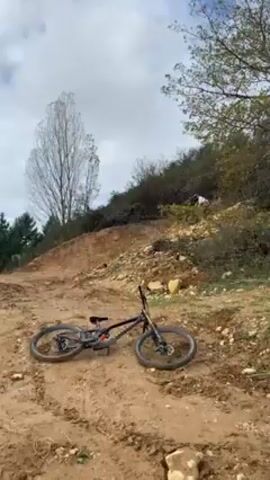 A person on a mountain bike rides downhill on a dirt trail with several jumps. The biker is wearing a white shirt, a helmet, and gloves. The trail is surrounded by trees and bushes. Another mountain bike is lying on the trail in the foreground. The sky is cloudy.
