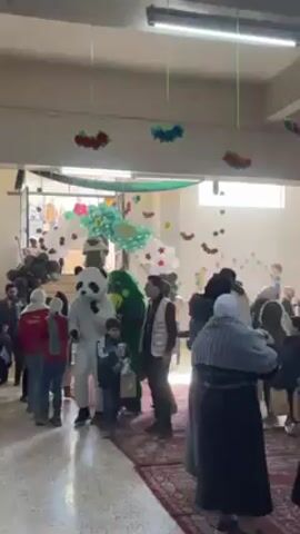The video shows a crowded indoor space, possibly a school hall, decorated with balloons and other decorations. A person in a panda costume walks through the crowd of people, mostly children and adults. Some adults wear vests with logos, suggesting they are volunteers or organizers. The children seem to be receiving gifts or items, as some hold bags. The overall atmosphere appears festive and cheerful.