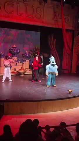 The video shows a stage performance featuring characters dressed as Aladdin characters. On the left, a character in a pink outfit is bent over, possibly a dancer. In the center, Jafar, in his iconic red and black outfit, wields a staff and appears to be interacting with Genie, who is dressed in blue and in a crouching position. To the right, another character in a dark outfit is visible, also in a crouching position. The stage backdrop features a large screen displaying an animated desert scene with a palace and a stormy sky, and to the right, a large decorative element resembling a golden swirl. Two small, golden lamps are placed on the stage floor. The stage is lit with red and pink lights, and the audience is visible in the foreground, silhouetted by the stage lighting.