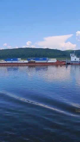 The video shows a barge being pushed by a tugboat on a river. The barge is loaded with shipping containers. The river is wide and calm, with ripples on the surface. In the background, there are trees and hills. The sky is blue with some white clouds. The tugboat has a white cabin and a grey hull with red trim. The barge is mostly red, with some blue and white containers on top. The video appears to be shot from another boat moving alongside the barge and tugboat.