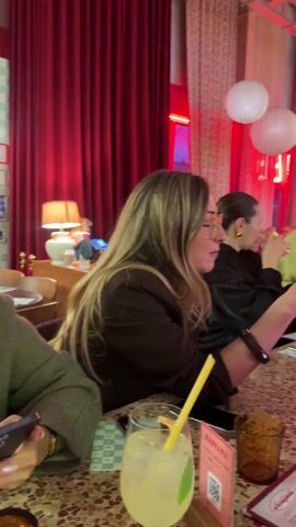 A group of people are seated at a table in a brightly lit restaurant with a modern decor. Red curtains and white spherical pendant lights dominate the scene. Several people are looking at the red menus. A woman with long blond hair laughs while looking at a menu, and others look on. Drinks with yellow straws are on the table, and other customers are visible in the background.