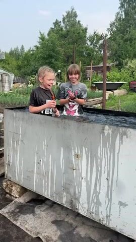Two children, a boy and a girl, are splashing in a large metal trough filled with water. The boy is on the left, wearing a black t-shirt. The girl is on the right, wearing a grey t-shirt with a cartoon character on it. They are both laughing and splashing water at each other. The trough is outdoors, with trees and a garden visible in the background. The trough appears to be old and rusted, with water stains running down the sides. There is a black liner inside the trough. The ground around the trough is wet and appears to be dirt or mud.