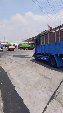A blue truck with a wooden cargo bed is seen from the rear as it turns right. The truck has a tarp covering the cargo. In the background, there is a gas station with a red roof and a white sign that reads "PERTAMINA". A red and white flag is flying from a pole at the gas station. There are other trucks in the distance, some green and some blue. The ground is paved with concrete, and there are some dark streaks on the pavement. The sky is blue with some white clouds. The scene appears to be an outdoor industrial area or a truck depot.