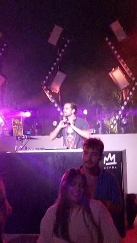 The video shows a nighttime outdoor party scene. A DJ, wearing a t-shirt with a graphic of a face and headphones, is standing behind a DJ booth, operating equipment. The booth has a logo that appears to be an "M" with wings, and the word "ALPHA" below it. In front of the booth, two people are dancing, a woman in a light-colored top and a man in a blue shirt. The environment is illuminated by bright purple and pink stage lights, with geometric lighting rigs visible above the DJ booth. The overall atmosphere suggests a lively music event.