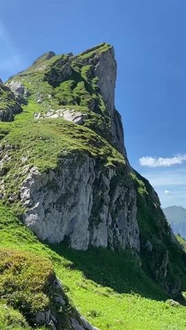 This is a vertical, slow pan video taken from a grassy mountainside looking out over a vast mountain range. The foreground features a steep, rocky cliff face covered in green grass. The middle ground shows more rolling green hills and a distant view of a valley with a bright turquoise lake and a small village nestled among the trees. The background is filled with layers of hazy, green mountains under a clear blue sky with scattered white clouds. The camera pans slowly from left to right, revealing more of the expansive landscape. There are no people or discernible animals visible in the video. The overall impression is one of natural beauty and serenity.