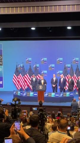 The video shows a press conference at what appears to be the NATO Summit in Ankara, based on the backdrop. In the foreground, the camera is pointed towards a stage where several people are standing behind a podium. The main speaker, identified as Donald Trump by his appearance, is at the podium. He is wearing a dark suit, a light blue shirt, and a yellow tie. He is speaking into a microphone. To his left, there are three American flags. Behind him, several other men in suits are standing, including one to his immediate left and two further back. The backdrop features the NATO logo and "ANKARA SUMMIT" in multiple locations. In the audience, many people are visible, holding up cell phones to record the event. There are also several cameras and media personnel in the audience area. The overall environment suggests a formal political event.