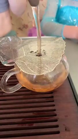 A close-up shot shows a clear glass teapot with orange tea in it. A decorative silver leaf-shaped strainer is placed on top of the teapot, and a clear liquid is being poured through it. The teapot sits on a wooden tray with slats. In the background, two people are visible, one wearing a blue top.