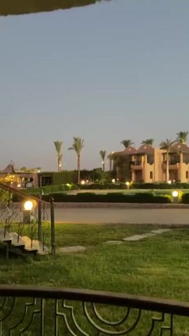 The video displays a twilight view of a resort or hotel complex. In the foreground, there is a well-maintained lawn with some stone pathways and a dark metal railing. A white bench is visible on the right side of the lawn. In the mid-ground, there is a road or pathway, and beyond that, a manicured garden area with several palm trees and bushes. The buildings in the background are light-colored, multi-story structures with arched windows and balconies, illuminated by warm outdoor lights. The sky is clear and pale blue, indicating dusk or dawn. The overall atmosphere is serene and peaceful, characteristic of a resort setting in the evening. No people or animals are clearly visible in the frame.