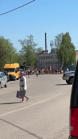The video shows a street with several runners participating in a race. There is a crowd of people gathered at the end of the street, near barricades, seemingly the finish line. Cars are parked on both sides of the street. A woman is crossing the street in the foreground. A yellow van and a silver SUV are also visible. The background includes a building with a tall chimney and trees lining the street. The sky is clear and blue.