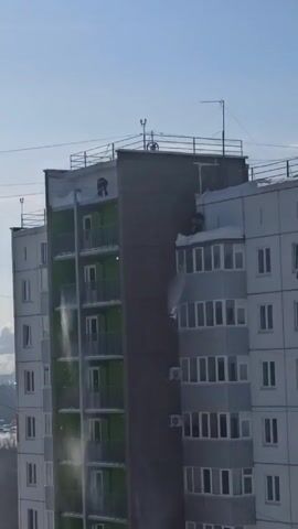 Two figures are on the roof of a tall, multi-story apartment building, which appears to be in a cold climate due to the presence of snow. The figures are actively shoveling snow off the edge of the building. The building has a distinctive color scheme, with some sections painted green. The sky is clear and blue, suggesting a sunny day. The surrounding environment is slightly hazy, with bare trees visible in the background.