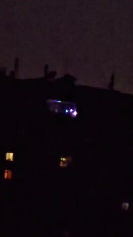 A dark, grainy video captures a nighttime cityscape with illuminated windows of buildings. The primary focus is a brightly colored light, possibly a reflection or a moving light source, that flashes red and then shifts to multi-colored hues. The scene appears to be in a residential area, with silhouetted rooftops and antennas visible against a dark sky. The time of day is clearly night, indicated by the darkness and the artificial lights. There are no identifiable people or specific objects, beyond the buildings and the mysterious light. The overall atmosphere is somewhat eerie due to the low visibility and the unidentifiable light source. The location context of Seleznevka, Kazakhstan, provides a geographical reference but doesn't offer specific visual cues within the short clip. No text is visible.