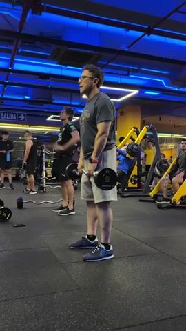 A man with glasses and a gray t-shirt is performing a barbell exercise in a gym. He is holding a barbell with weights on each end and is in the process of lifting it to his chest, with his arms bent at the elbows. He is wearing light-colored shorts and blue and white athletic shoes. Other people are visible in the background, some using gym equipment, and the gym has blue lighting overhead.
