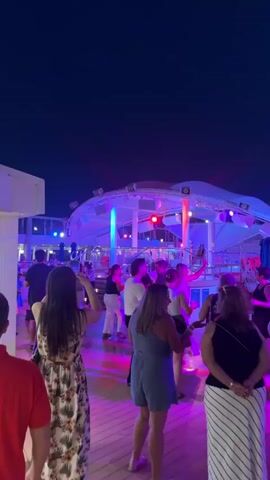 This is a vertical video of a crowded outdoor dance floor at night.  The dance floor is lit by colorful stage lights, primarily pink and blue, casting vibrant reflections. A DJ booth is visible in the background on a raised platform with white structures and more lights. People of various ages are dancing and socializing. In the foreground, several women are facing away from the camera, watching the dancers. One woman in a floral dress is holding her phone up, possibly recording. Others are standing or dancing. The overall atmosphere is lively and celebratory.