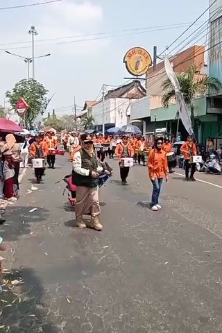 A street parade is underway. A woman in a tactical vest and a baseball cap walks in the foreground, facing the camera. Behind her, a group of people in matching orange shirts march in formation. Some are carrying drums. Spectators line the sides of the street. Buildings are visible on the right side of the street, and trees and a utility pole are on the left. The sky is bright and clear.