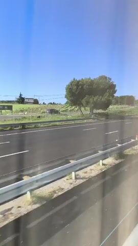 From a moving vehicle, the video shows a highway on a bright, clear day. The highway has multiple lanes and a median barrier. Various vehicles, including cars and vans, are traveling along the highway. Beyond the highway are grassy fields, scattered trees, and utility poles, all under a clear blue sky. A road sign indicates the highway number as "A709" and a distance "9.506". The perspective is from within another vehicle, looking out through a window.