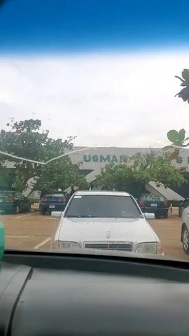 The video shows the view from inside a car in a parking lot. Visible through the windshield are several parked cars, including a white Mercedes-Benz and a silver Toyota Camry. In the background, the facade of a building labeled "USMAN" is partially obscured by trees. A box of tissues with a deer decoration sits on the dashboard of the car. The sky is overcast, and the camera is relatively stable.