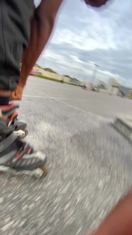 The video shows a person wearing inline skates and black pants on an asphalt surface. The camera is positioned low to the ground, focusing on the skates as the person moves forward. The skates are gray and black with silver frames and worn wheels. The asphalt has a mottled gray texture. In the background, there's a blurry view of a parking lot, buildings, and a red gas station. The sky is overcast. The camera angle is unstable, creating a spinning effect as the person skates.
