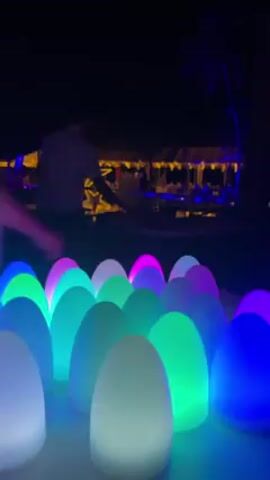 The video shows a dimly lit outdoor scene at night, focused on a collection of oval-shaped lights in various colors (blue, green, pink, white). In the background, out of focus, are people walking and a building with lights visible. There are tables visible as well with unknown items. The overall atmosphere seems festive or decorative due to the colorful lights.