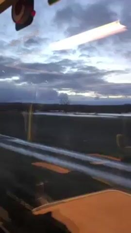 The view from inside a moving train reveals a landscape outside. The cloudy sky is a mix of gray and blue, with hints of sunset glow. The ground is mostly flat and appears to be covered with water or flooded. A few trees can be seen in the distance. The window reflects the interior of the train, including a safety instruction sticker. A light fixture above is also visible. The train is moving, blurring the foreground.
