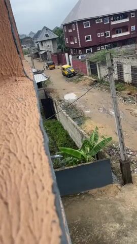 This video shows an outdoor scene with a dirt road. Two yellow vehicles, likely auto-rickshaws, are visible on the road. One is further away, and the other is closer to the foreground. The environment appears somewhat unkempt, with some debris visible and evidence of puddles on the ground, suggesting recent rain or a poorly drained area. Buildings line the sides of the road, with varying architectural styles. A large, dark orange wall is prominently in the foreground on the left side of the frame, obscuring part of the view. A utility pole with wires is also visible. There are no people clearly visible in the frame interacting with the vehicles or the environment.