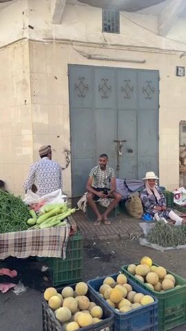 The video depicts an outdoor market scene with three people sitting near produce stands. To the left, a man in a patterned shirt stands behind a table covered with green vegetables and zucchini. A man in a patterned shirt and shorts sits on a crate in front of a large, closed metal door. Next to him, a woman wearing a hat and patterned clothing sits on the ground amidst piles of herbs. Several crates filled with melons are in the foreground. The setting is an urban street with buildings showing signs of age. There's graffiti on the wall to the right, and people are seen passing by in the background.