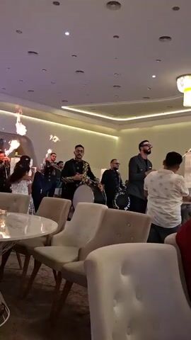 A group of men in ornate dark jackets and pants are walking in a room. One man in the front is playing a drum and another is holding a microphone. There is also a man holding a flaming torch in the air. Several people are seated at tables in the foreground, some of whom are looking at the performers. The room has white walls and a white ceiling with recessed lighting.