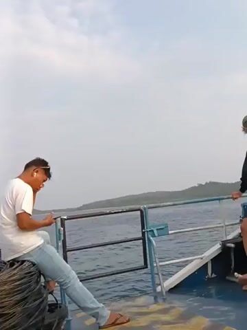 Two men are on the deck of a boat. The man on the left is wearing a white t-shirt, blue jeans, and has dark hair. He is sitting down and looking at his phone. The man on the right is wearing a black hoodie, black shorts, a baseball cap, and glasses. He is standing and looking out at the water. There are railings on the boat. The water is calm and there are islands in the distance. The sky is overcast.