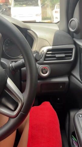 A person wearing a red garment is sitting in the driver's seat of a car. Their right hand, adorned with white nail polish, a gold ring, and a gold bracelet, reaches towards the car's start button. The hand presses the start button, which illuminates with the word "ENGINE START STOP." The car dashboard and steering wheel are visible, and the video is taken from an interior perspective. A white van is visible through the windshield.