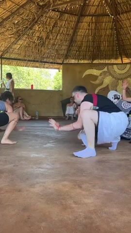 This video captures a group of people engaged in what appears to be a dance or movement session within a circular, open-air structure with a thatched roof. The ground is a textured, earthy surface. Several individuals are visible in the foreground and mid-ground. On the left, a woman wearing black shorts and a grey tank top is in a low, quadrupedal stance, facing away from the camera. To her right, another person, facing away from the camera, wears white shorts, a black t-shirt with red suspenders, and light purple socks, also in a crouching position. Their left arm is extended, and thin red threads are visible around their wrist and extending towards another person. In the mid-right, a person wearing a patterned dress and headscarf is also in a crouching or kneeling position. In the background, several other individuals are seated or standing. One person in a white dress stands near a window-like opening, holding a white object. The overall atmosphere suggests a communal activity or workshop.