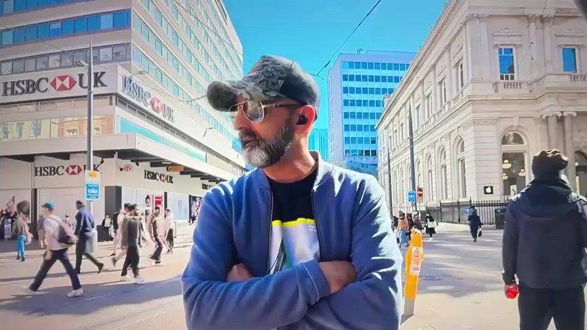A man wearing a camouflage baseball cap, sunglasses, and a grey jacket stands with his arms crossed, looking to his left. He has a beard and appears to be wearing earbuds. The background shows a street scene with people walking and buildings, including one with an HSBC UK sign. The sky is blue and sunny. The man is positioned in the foreground, slightly off-center, with other people and the urban environment visible behind him.